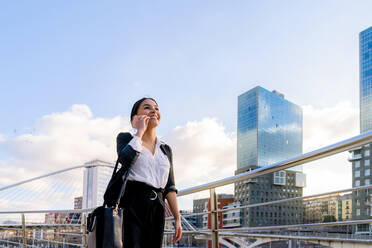 Cheerful young ethnic female entrepreneur talking on cellphone while looking away against city river in sunlight - ADSF26586