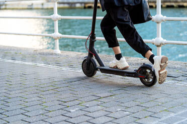 Seitenansicht der Ernte anonyme Frau in Turnschuhen Reiten modernen Roller auf dem Gehweg gegen Stadt Fluss - ADSF26589