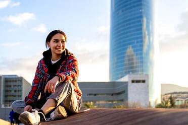 Happy ethnic female sitting with crossed legs in town while looking away in sunlight - ADSF26599