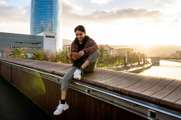 Young smiling ethnic female in casual wear text messaging on cellphone on bridge over river under cloudy sky in sunlight - ADSF26601