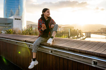 Young smiling ethnic female in casual wear text messaging on cellphone on bridge over river under cloudy sky in sunlight looking away - ADSF26602