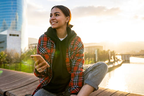Junge lächelnde ethnische Frau in Freizeitkleidung Textnachrichten auf Handy auf Brücke über Fluss unter bewölktem Himmel im Sonnenlicht wegschauen - ADSF26603