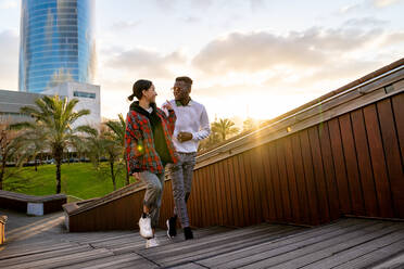 Cheerful diverse multiethnic couple in stylish apparel speaking while looking at each other and strolling on city staircase in sunlight - ADSF26604