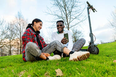 From below of cheerful multiracial couple in trendy wear with cellphone speaking on meadow with electric scooter in daytime - ADSF26608