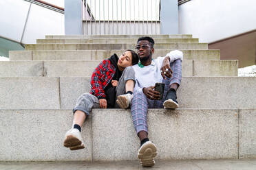 Junges gemischtrassiges Paar in trendiger Kleidung sitzt auf einer Treppe in der Stadt mit einem Smartphone und schaut weg - ADSF26611