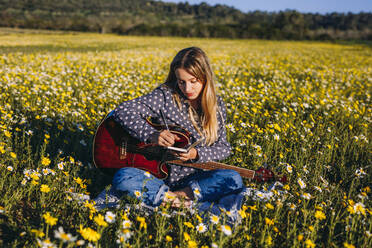 Young hipster woman sitting on a meadow in the countryside writing songs on notebook and playing guitar during summer sunlight. - ADSF26612