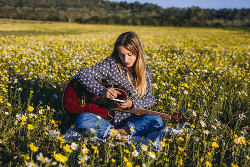 Junge Hipster-Frau sitzt auf einer Wiese auf dem Land und schreibt Lieder in ihr Notizbuch und spielt Gitarre in der Sommersonne. - ADSF26612