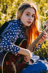 Nachdenkliche junge Hipster-Frau, die auf einer Wiese in der Natur sitzt, Lieder in ein Notizbuch schreibt und bei sommerlichem Sonnenlicht Gitarre spielt - ADSF26613