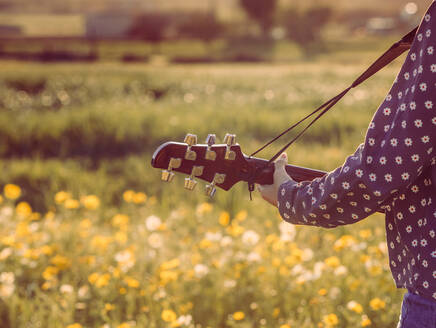 Rückenansicht einer abgeschnittenen, nicht erkennbaren jungen Hipster-Frau, die auf einer Wiese im Grünen steht und bei sommerlichem Sonnenlicht Gitarre spielt - ADSF26614
