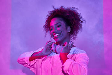 Fröhliches Porträt einer jungen Afroamerikanerin in weißer Kleidung, die Kopfhörer im Nacken trägt und in die Kamera schaut, während sie in einem dunklen Studio mit Neonlicht steht - ADSF26622