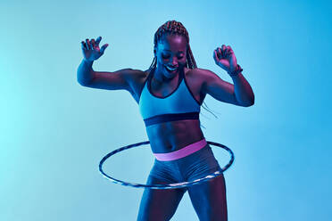 Cheerful African American sportswoman in activewear rotating hoop during training while looking down in neon light - ADSF26647