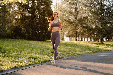 Fröhliche Sportlerin in Sportkleidung, die auf einer asphaltierten Straße läuft, während sie beim Ausdauertraining im Sonnenlicht wegschaut - ADSF26659