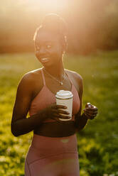 Zufriedene afroamerikanische Sportlerin in Sportkleidung, die abends mit einer Tasse Getränk im Park steht und wegschaut - ADSF26663