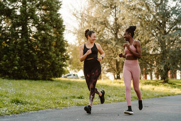 Lächelnde, gemischtrassige Läuferinnen in Sportkleidung joggen und unterhalten sich beim Ausdauertraining auf einem Gehweg in der Stadt - ADSF26665