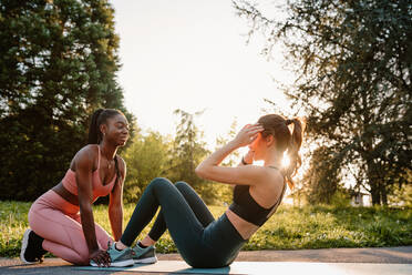 Seitenansicht einer schlanken Sportlerin, die mit Hilfe einer schwarzen Sportlerin während des Trainings bei Sonnenuntergang im Park Bauchmuskeln trainiert - ADSF26666