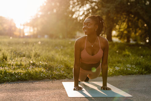 Fitte afroamerikanische Sportlerin balanciert in der Plank-Position, während sie im Park bei Sonnenuntergang Bauchmuskeltraining macht - ADSF26668