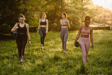 Selbstbewusste, fitte, multirassische Sportlerinnen, die mit einer Matte auf dem Rasen laufen, während sie im Sommer im Park trainieren (abends) - ADSF26669