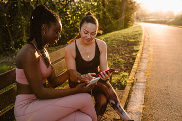Fröhliche, gemischtrassige Sportlerinnen in Sportkleidung, die nach dem Training bei Sonnenuntergang auf einer Bank im Park sitzen und gemeinsam Mobiltelefone benutzen - ADSF26670