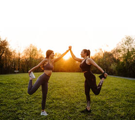 Seitenansicht von lächelnden sportlichen Läuferinnen, die sich an den Händen halten und die Beine strecken, während sie sich gegenseitig beim Training im Park bei Sonnenuntergang unterstützen - ADSF26674