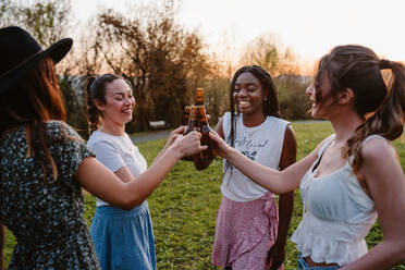 Eine Gruppe glücklicher Frauen versammelt sich im Park und stößt mit Bierflaschen an, während sie ein Sommerwochenende zusammen genießen - ADSF26676