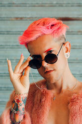 Young gay in stylish wear with tattoo, pink hair and manicure with fluffy jacket holding sunglasses while looking at camera - ADSF26681