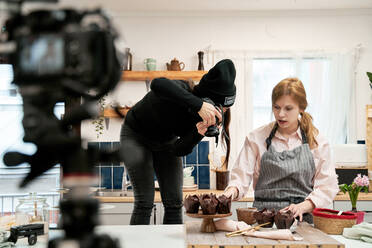 Unrecognizable woman taking photo of chocolate muffins on digital camera against blogger talking during cooking process in kitchen - ADSF26685