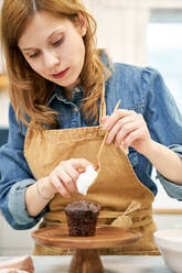 Smiling young female in apron with sweet cream on whisk looking down during cooking process at home - ADSF26688