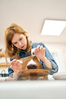 Eine junge Frau dekoriert einen Schokoladenkuchen mit süßer Schlagsahne auf einem Kuchenständer, während sie zu Hause kocht - ADSF26689