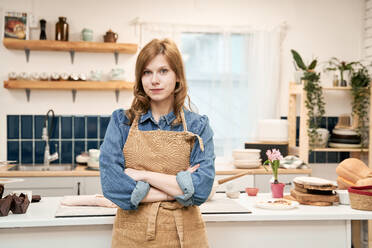 Young self assured female with folded arms in apron looking at camera at home - ADSF26690