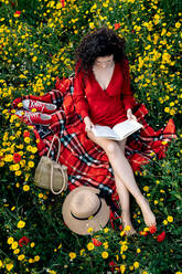 From above of barefoot female with crossed legs reading textbook on blanket with hat and handbag among blossoming daisies - ADSF26700