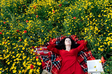Von oben auf eine Frau mit geschlossenen Augen, die auf einer Decke mit einer Handtasche zwischen blühenden Gänseblümchen an einem Frühlingssommertag eine Lesepause einlegt - ADSF26702