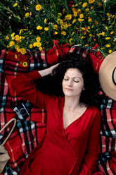 Draufsicht auf eine zarte Frau mit geschlossenen Augen, die ihr Haar berührt, während sie auf einer karierten Decke liegt, mit einer alten Fotokamera im Feld - ADSF26706