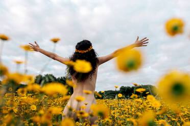 Back view of anonymous naked brunette in flower wreath enjoying meadow with blossoming daisies under cloudy sky in summer - ADSF26709