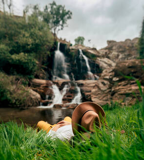 Anonymer männlicher Reisender mit Hut, der sich am Ufer eines üppigen Sees entspannt und die Aussicht auf einen reißenden Wasserfall in der sommerlichen Natur genießt - ADSF26711