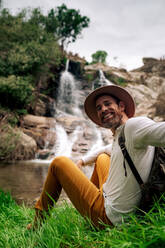 Fröhlicher erwachsener männlicher Wanderer in Freizeitkleidung mit Rucksack am Seeufer in der Nähe eines malerischen Wasserfalls sitzend, während er die Kamera berührt und anschaut - ADSF26712