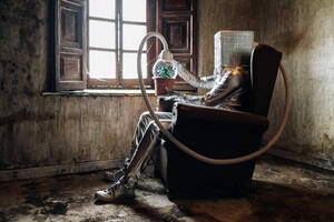 Seitenansicht unerkennbare Person in silbernem Kostüm mit Atemgerät und Schlauch, der an einer Topfpflanze befestigt ist, die auf einem Stuhl in einem verwitterten, verlassenen Haus sitzt - ADSF26716