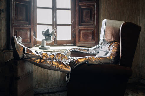 Side view unrecognizable person wearing protective silver suit with box on head resting on armchair in shabby room in abandoned house - ADSF26719