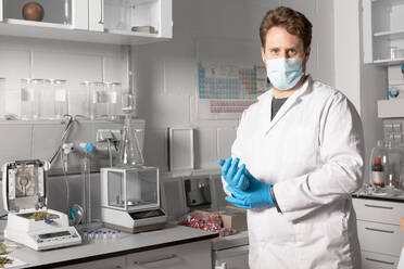 Male chemist in sterile mask and gloves looking at camera against moisture analyzer and analytical balance in cannabis laboratory - ADSF26729