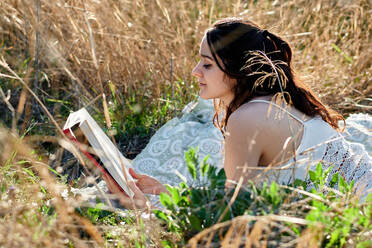 Dreamy charming brunette in white dress lying on field meadow and reading book in sunlight - ADSF26760