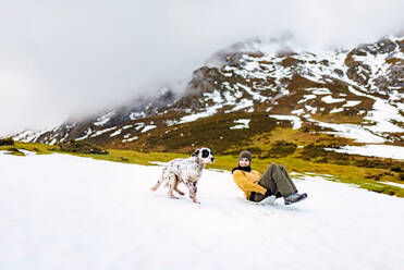 Happy woman sliding down slope covered with snow while happy English Setter dog running aside in mountains - ADSF26780