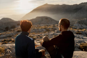 Back view anonymous male friends in casual wear resting on rocky mountain summit and admiring scenic sunset over rough highlands in Seville Spain - ADSF26782