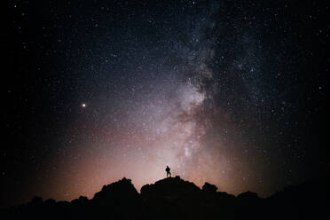 Silhouette of anonymous tourist standing on cliff against glowing starry sky at night - ADSF26787