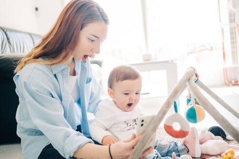 Playful shocked woman showing mobile phone to charming toddler while spending time at home - ADSF26791