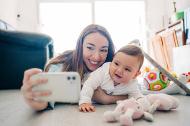 Verspielte Frau, die ein Selfie mit einem bezaubernden Kleinkind auf dem Mobiltelefon macht, während sie Zeit zu Hause verbringt - ADSF26793