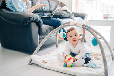 Abgeschnittene, nicht erkennbare Mutter in Freizeitkleidung, die auf einem Smartphone und Netbook surft und auf dem Sofa neben einem entzückenden kleinen Baby sitzt, das in die Kamera schaut, während es mit Spielzeug auf dem Boden im Wohnzimmer spielt - ADSF26795