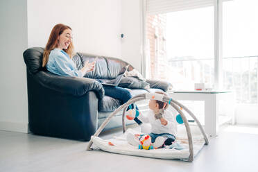 Fokussierte junge Mutter in legerem Outfit, die mit Smartphone und Netbook auf dem Sofa sitzt, in der Nähe eines entzückenden kleinen Babys, das mit Spielzeug auf dem Boden im Wohnzimmer spielt - ADSF26796