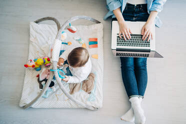 Top view anonymous mother wearing casual outfit sitting on floor with netbook on laps and outstretching hand to cute little baby playing with toys on floor - ADSF26797