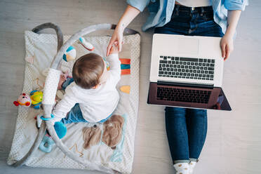 Top view anonymous mother wearing casual outfit sitting on floor with netbook on laps and outstretching hand to cute little baby playing with toys on floor - ADSF26798