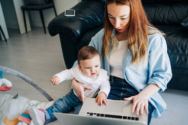 Junge Mutter in Freizeitkleidung, die ihr süßes Baby umarmt und auf dem Boden sitzend auf ihrem Netbook surft - ADSF26799