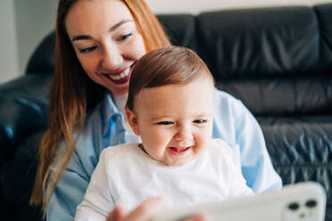 Glückliche junge Mutter und entzückendes lächelndes Baby, das sich ein lustiges Video auf dem Handy ansieht, während es auf dem gemütlichen Sofa zu Hause sitzt - ADSF26801